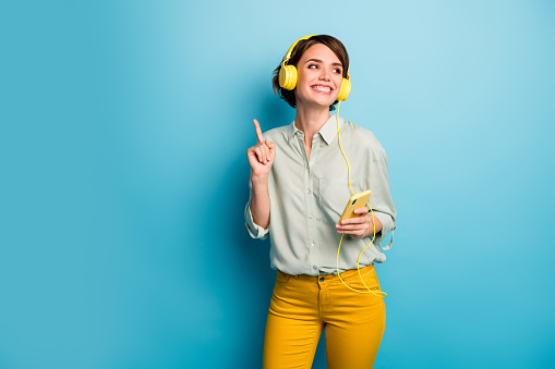 Photo of funny cheerful lady hold telephone listening radio cool stylish, earphones dancing raise finger wear casual green shirt yellow trousers isolated blue color background