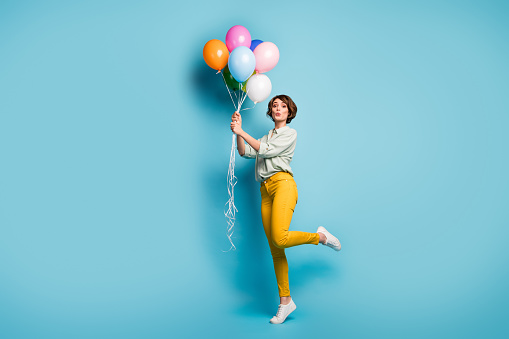 Full size profile photo of funny lady surprise birthday party hold many air balloons playful weekend mood wear casual green shirt yellow pants footwear isolated blue color background