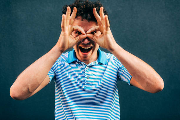 istock Funny young man posing for advertisement, smiling and making a grimace, standing at building gray wall outdoors. Happy student male has positive expression, pretends to wear binocular. 1210314742