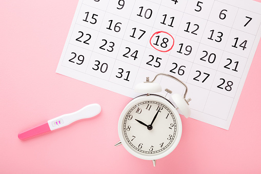 Calendario, prueba con dos rayas y reloj despertador blanco sobre fondo de mesa rosa pastel. Resultado positivo. Comienza el embarazo. Closeup. Vista de arriba hacia abajo. photo