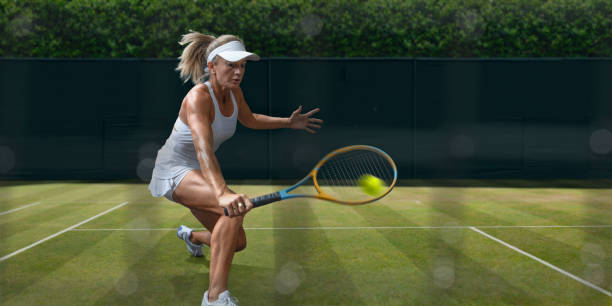 Female Tennis Player In Mid Motion During Grass Court Game A female tennis player dressed in a white tennis outfit including visor, leaping in mid-motion, with arm holding racket outstretched as she hits the ball in a volley. The player is viewed through the net and is playing on a grass court with fence and hedge in background. backhand stroke stock pictures, royalty-free photos & images