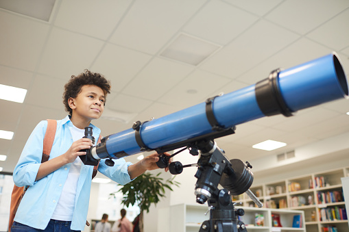 Horizontal medium low angle shot of African American middle schooler using modern telescope to at something far away, copy space