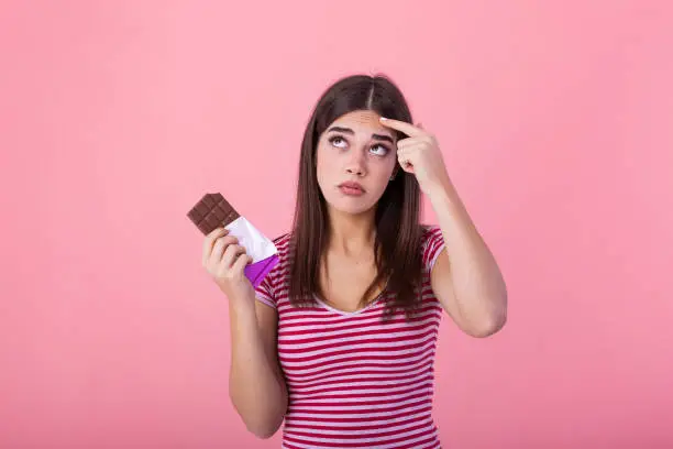 Teenage girl with acne problem holding chocolate bar against pink background. Young beautiful Woman Acne Problem Face with Chocolate bar UnHappy Eating,His Stressful face.