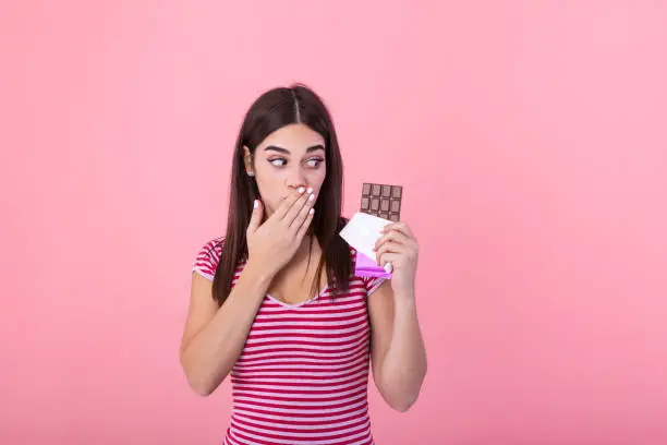 Photo of Portrait of a happy young woman with chocolate bar isolated over pink background covenring her mouth. Young woman with natural make up having fun and eating chocolate isolated on pink background
