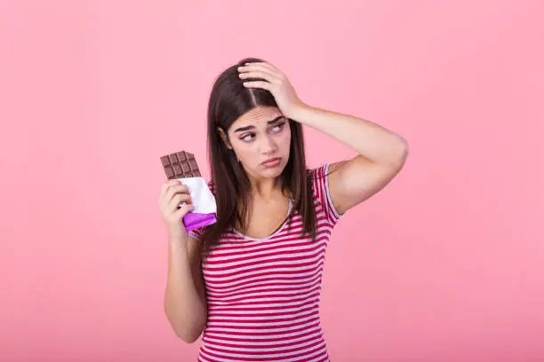 Photo of Portrait sad young woman tired of diet restrictions craving sweets chocolate isolated on pink wall background. Human face expression emotion. Nutrition concept. Feeling of guilt. Passion for chocolate