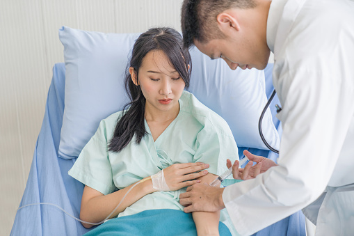 Doctor or nurse gives vaccine or medicine injection to adult Asian descent patient in office or clinic setting. She uses syringe and wears gloves. Medical exam, consultation, therapist.