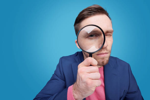 Serious young male detective holding magnifying glass by right eye Serious young male detective holding magnifying glass by right eye while standing in front of camera in isolation looking through an object stock pictures, royalty-free photos & images