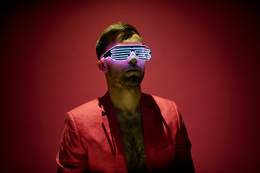Contemporary young man in red jacket and high tech eyewear standing in darkness against maroon background in isolation