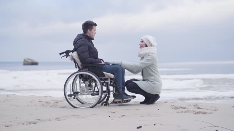 Young woman sitting on hunkers in front of boy in wheelchair and talking. Mother or invalid tender supporting handicapped young man on sea shore or river bank outdoors. Joy, lifestyle, help.