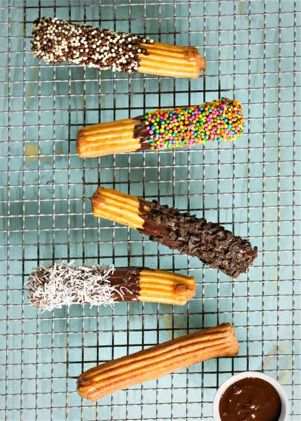 Five delicious churros on a grill, with different types of coverage, such as chocolate sprinkle, pieces of cookies, grated coconut and melted chocolate.
