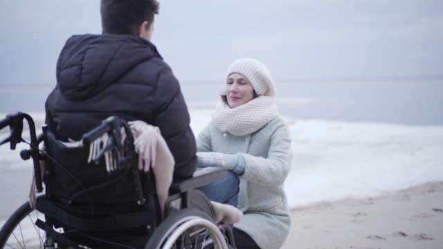 Portrait of strong adult Caucasian woman talking with man in wheelchair outdoors. Positive mother or invalid tender sitting on hunkers in front of son or patient at river bank. Support, disability.