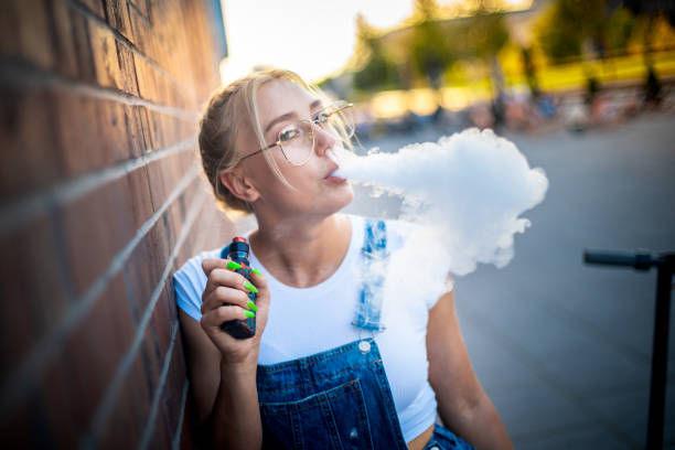 современная девушка на открытом воздухе курение вейпинг электронная сигарета делает большое облако дыма - quit scene стоковые фото и изображения
