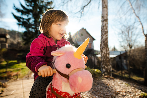 Charming playful girl sitting on a fluffy unicorn toy and riding it in her backyard