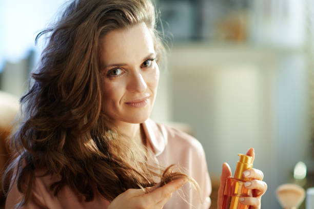 modern woman holding hair oil bottle and checking hair ends Portrait of modern middle age woman in pajamas with long brunette wavy hair holding hair oil bottle and checking hair ends in the modern living room in sunny winter day. hair care women mature adult human skin stock pictures, royalty-free photos & images