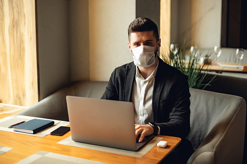 Adult bearded man indoors in cafe. Lifestyle concept photo with copy space. Picture of professional with disposable protective face mask and gray personal computer