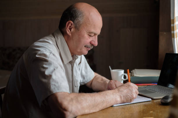 istock Senior man is writing in his room sitting near window. 1210356494