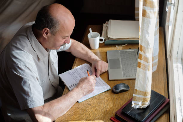 istock Senior man is writing in his room sitting near window. 1210357007