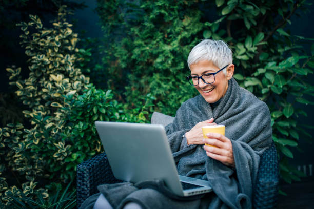 mujer mayor sentada en una silla en el jardín del patio trasero y mirando la computadora portátil, retrato. - red chairs fotografías e imágenes de stock
