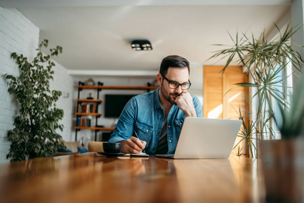 homem sério olhando para o laptop e tomando notas em notebook, retrato. - household finances - fotografias e filmes do acervo