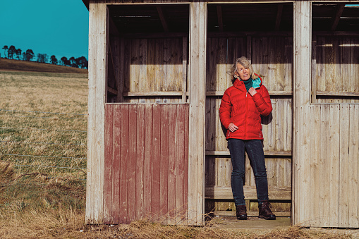 Mature woman in red padded jacket waiting at an old wooden bus stop in a country location.