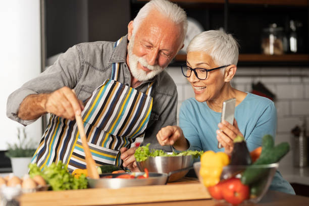 pareja de la tercera edad comparte una videollamada con sus amigos cómo preparar la comida - cooking senior adult healthy lifestyle couple fotografías e imágenes de stock