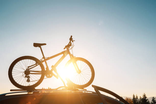 istock Mounted mountain bicycle silhouette on the car roof with evening sun light rays background. Safe sport items transportation using a car concept image. 1210361894
