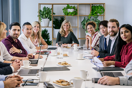 Wide angle view of ethnically diverse male and female business associates sitting at board room conference table and smiling at camera.