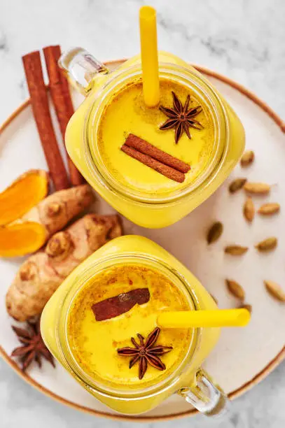 Photo of Hadi Doodh know as Golden Turmeric Milk in Mason Jar on white marble background. Golden Milk is ayurvedic recipe of hot milk with turmeric, ginger and spices.