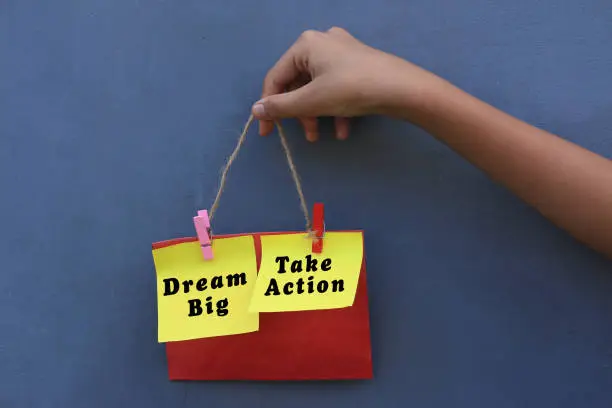 Inspirational quote - Dream big. Take action. With young girl holding origami paper notes rope hanging on wall. Motivational business typography words concept with colorful creative papers.