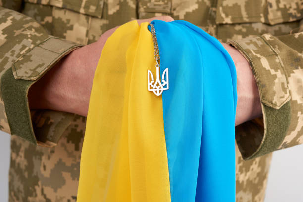 Ukrainian man warrior dressed in a military pixel uniform holds the yellow-blue flag of the state of Ukraine and on the chain a small coat of arms Ukrainian man warrior dressed in a military pixel uniform holds the yellow-blue flag of the state of Ukraine and on the chain a small coat of arms of the country of trident, close up khaki green photos stock pictures, royalty-free photos & images