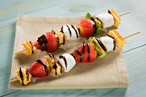 Two skewers with strawberrys, marshmallows and churros, covered with melted chocolate. A delicious dessert served on a bege napkin on a blue wooden table.