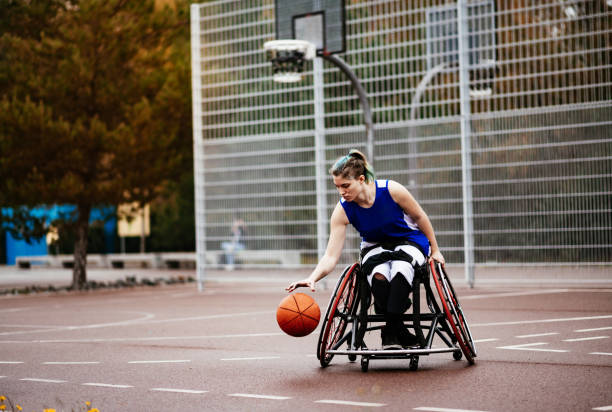 Woman in wheelchair playing basketball Motivated wheelchair basketball player shooting on the basketball court outdoors on bad weather day. leanincollection stock pictures, royalty-free photos & images