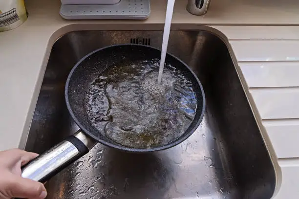 POV of frying pan with non-stick surface and hot oil under water tap flow in sink. Care kitchenware concept