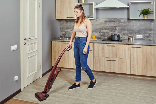 Caucasian woman uses a vacuum cleaner in kitchen. A young Caucasian woman vacuums a kitchen floor using a cordless vertical vacuum cleaner, or electric broom. The maid is cleaning up the hostels cook room. One white female do cleanup of indoors. cordless phone stock pictures, royalty-free photos & images