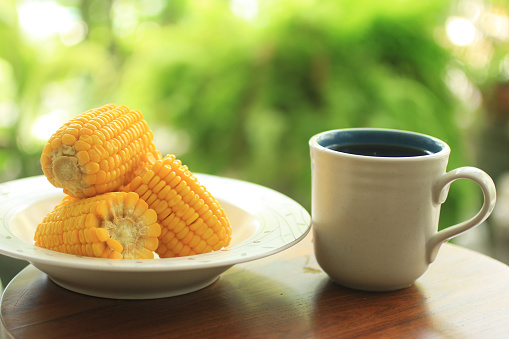 Boiled sweet corns on white plate and morning tea cup on the table. With blur green bokeh background.