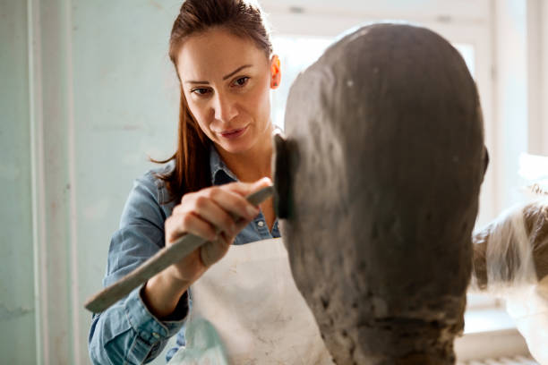 istock Young woman working on a head sculpture out of clay in workshop using tools for clay shading 1210417882