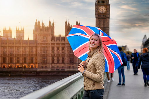 A blonde tourist woman with a British flag umbrella stands in front of the Big Ben Tower in London, Westminster, during a sightseeing trip through the city, United Kingdom