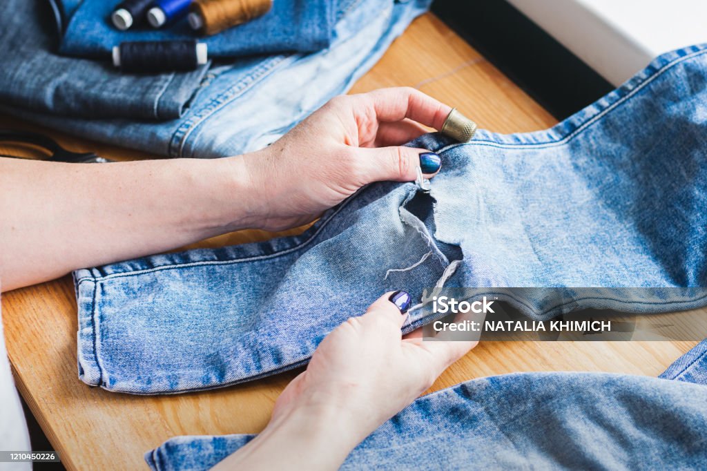 A woman is repairing ripped blue jeans. The concept of economical reuse of things. Homemade needlework. A woman is repairing ripped blue jeans. The concept of economical reuse of things. Homemade needlework. Horizontal frame selective focus Clothing Stock Photo
