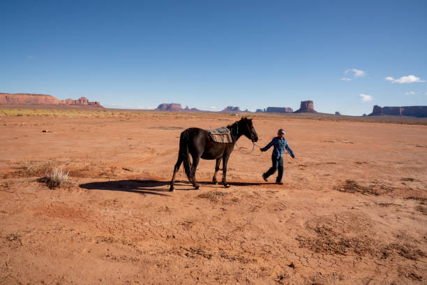 junge navajo geschwister auf ihren pferden mit einer felsformation hinter ihnen - monument valley navajo mesa monument valley tribal park stock-fotos und bilder