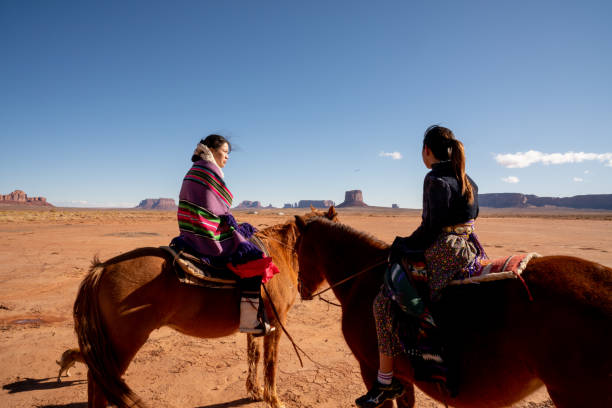 junge navajo geschwister auf ihren pferden mit einer felsformation hinter ihnen - monument valley navajo mesa monument valley tribal park stock-fotos und bilder