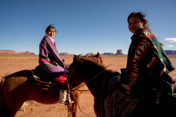 junge navajo geschwister auf ihren pferden mit einer felsformation hinter ihnen - monument valley navajo mesa monument valley tribal park stock-fotos und bilder