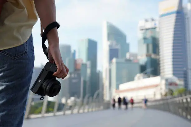 Photo of Close-up hand holding camera of  woman traveler with backpack and hat traveling into Singapore city downtown. Travelling in Singapore concept.