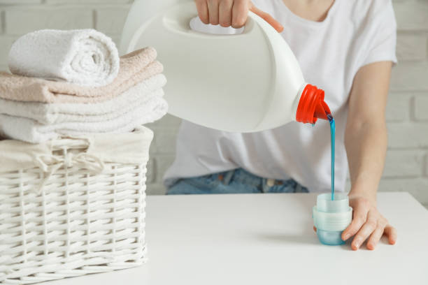 istock Close up of female hands pouring liquid laundry detergent into cap on white rustic table with towels on background in bathroom 1210484176