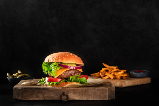 istock Burger with beef, cheese, onion, tomato, and green salad, a side view on a dark background with French fries and pickles, with a place for text 1210485460