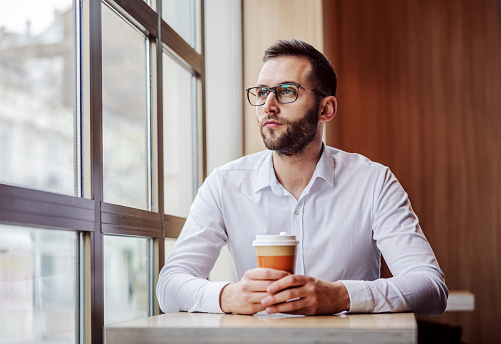 Young thoughtful man elegantly dressed sitting in fast food restaurant, holding disposable cup of coffee and looking trough window. He is having lunch break.