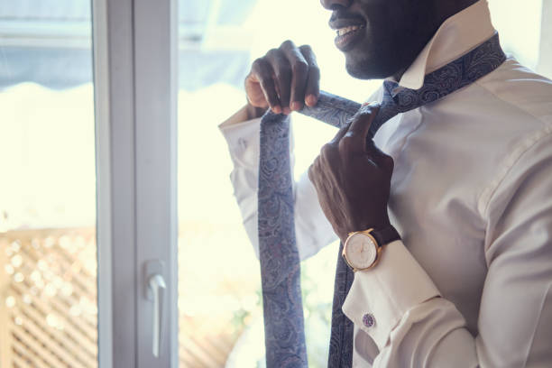 Elegant man is dressing up near window Smart american man is dressing up tie by himself while standing near window. necktie businessman collar tied knot stock pictures, royalty-free photos & images