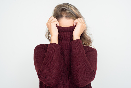 Woman hiding face in turtleneck. Young blonde woman wearing dark red knitted sweater and hiding head in maroon collar. Unrecognizable person concept