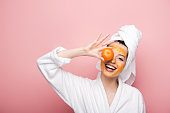 cheerful girl with citrus facial mask covering eye with tangerine on pink background