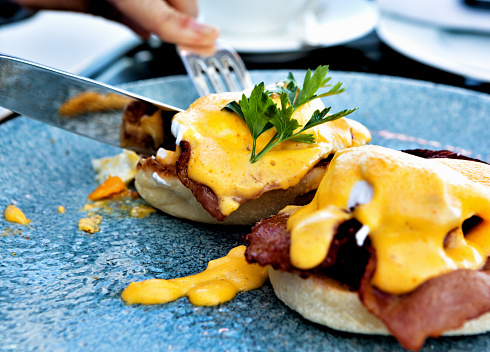 A knife and fork slices into a luxurious and delicious looking plate of Eggs Benedict.