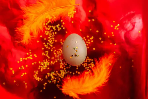 A chic festive pure Easter egg with star spangles with yellow feathers around it on a red mettalic shiny three-dimensional background. Happy Easter art Deco style, flat lay